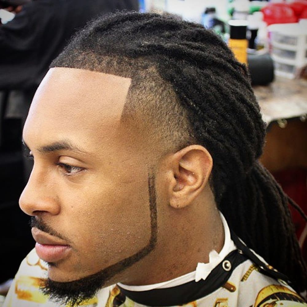 A man with neatly styled dreadlocks and a chin strap beard style. The beard is sharply trimmed, creating a clean, angular outline along the jawline and connecting with a precise mustache. The haircut features a fade on the sides, accentuating the contrast between the beard and the hairstyle. The overall look is sharp, well-groomed, and modern, emphasizing facial features.
