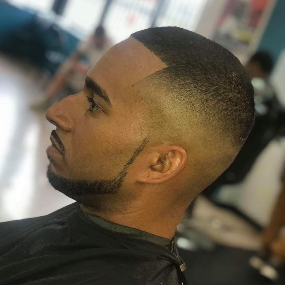 A man with a faded buzz cut hairstyle featuring a sharp, clean hairline and a neatly groomed chin strap beard style. The beard is precisely trimmed, outlining his jawline and connecting to a short, defined mustache, adding sharpness to his overall look. The smooth fade on the sides complements the clean angles of the beard, creating a polished and modern appearance. The background suggests a barber shop setting, emphasizing the professional grooming.