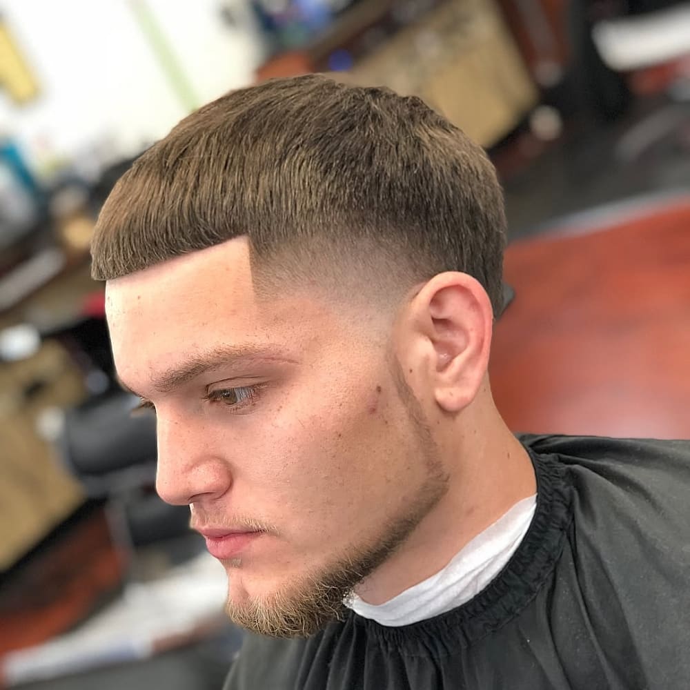 A young man showcasing a clean, modern haircut with a sharp fade on the sides and a textured crop on top. He has a chin strap beard style, precisely trimmed to outline his jawline and accentuate his facial structure. The beard connects seamlessly with a neatly groomed goatee, adding balance to his look. The overall grooming highlights attention to detail and a contemporary style.