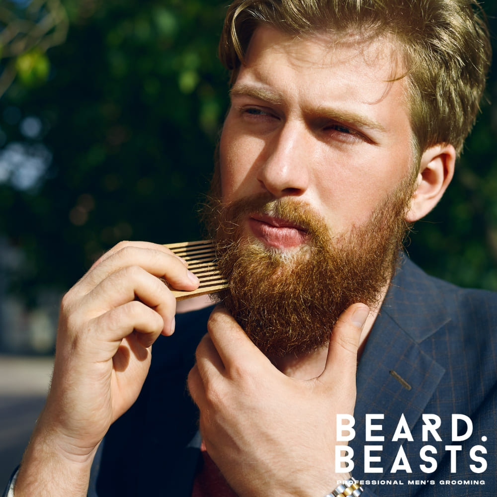 Outdoor portrait of a well-groomed man with a red beard, combing his facial hair with a wooden beard comb. Dressed in a suit, he exudes confidence and style.