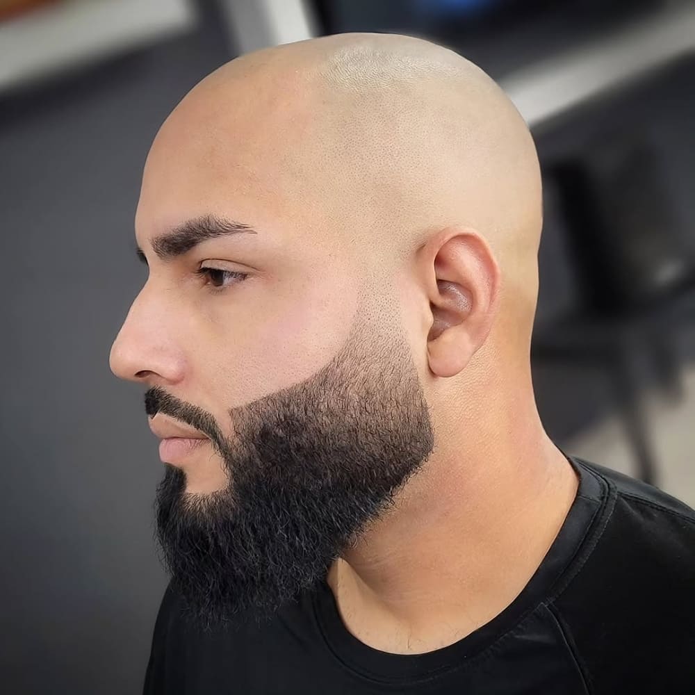 A well-groomed beard with faded sides on a man with a triangle face shape, featuring a wide jawline and a narrow forehead. The beard has a precise, gradual fade that blends seamlessly into the sideburns, enhancing facial symmetry. The sharp cheek lines and structured jawline create a clean, modern look, ideal for balancing strong facial angles. His bald head and dark, full beard add contrast, making this style stand out. The