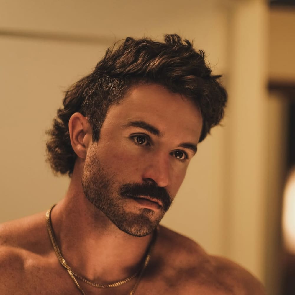 A stylish man with a beardstache, featuring a thick, well-groomed mustache and short stubble beard, ideal for a triangle face shape with a wide jawline and narrow forehead. The bold mustache draws attention upward, helping to balance facial proportions, while the light stubble softens the jawline. His textured mullet hairstyle adds to the rugged, masculine aesthetic. The warm indoor lighting and gold chains enhance the vintage, confident look, making this a perfect example of a beardstache for a triangle face shape.