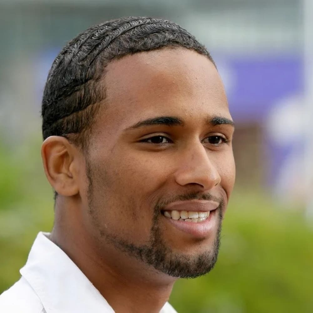 A young man with a neatly groomed beard and a chin strap with a circle beard combination smiles in an outdoor setting. His facial hair is well-defined, with a thin, precise chin strap extending along his jawline and connecting to a rounded goatee. He has a clean-shaven upper cheek area, highlighting the sharp contrast between his beard and skin. His hair is styled in short, wavy waves, complementing his polished look.