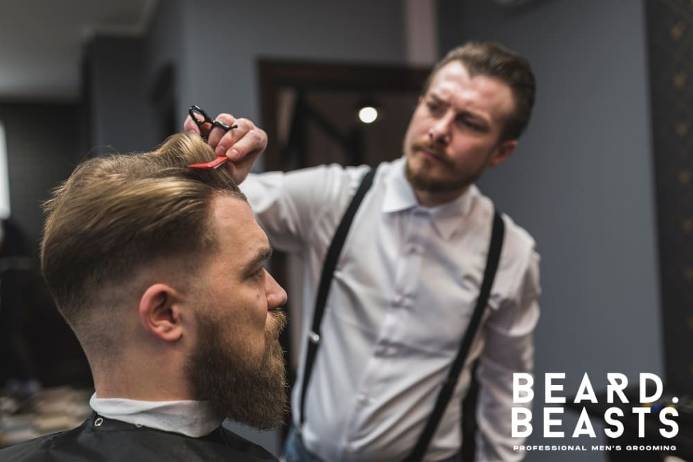 A professional barber, dressed in a white shirt with black suspenders, carefully trims a client's hair using scissors and a comb in a stylish barbershop. The client, a bearded man, sits in a barber chair with a fresh undercut hairstyle, featuring a neatly faded side and a longer, slicked-back top. The scene captures the attention to detail and craftsmanship of traditional men's grooming.