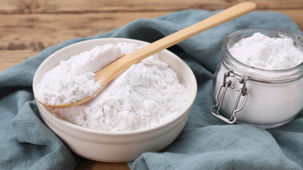 A bowl of baking soda with a wooden spoon, next to a glass jar filled with more baking soda. A natural remedy that can help with how to get pomade out of your hair by breaking down buildup and residue.