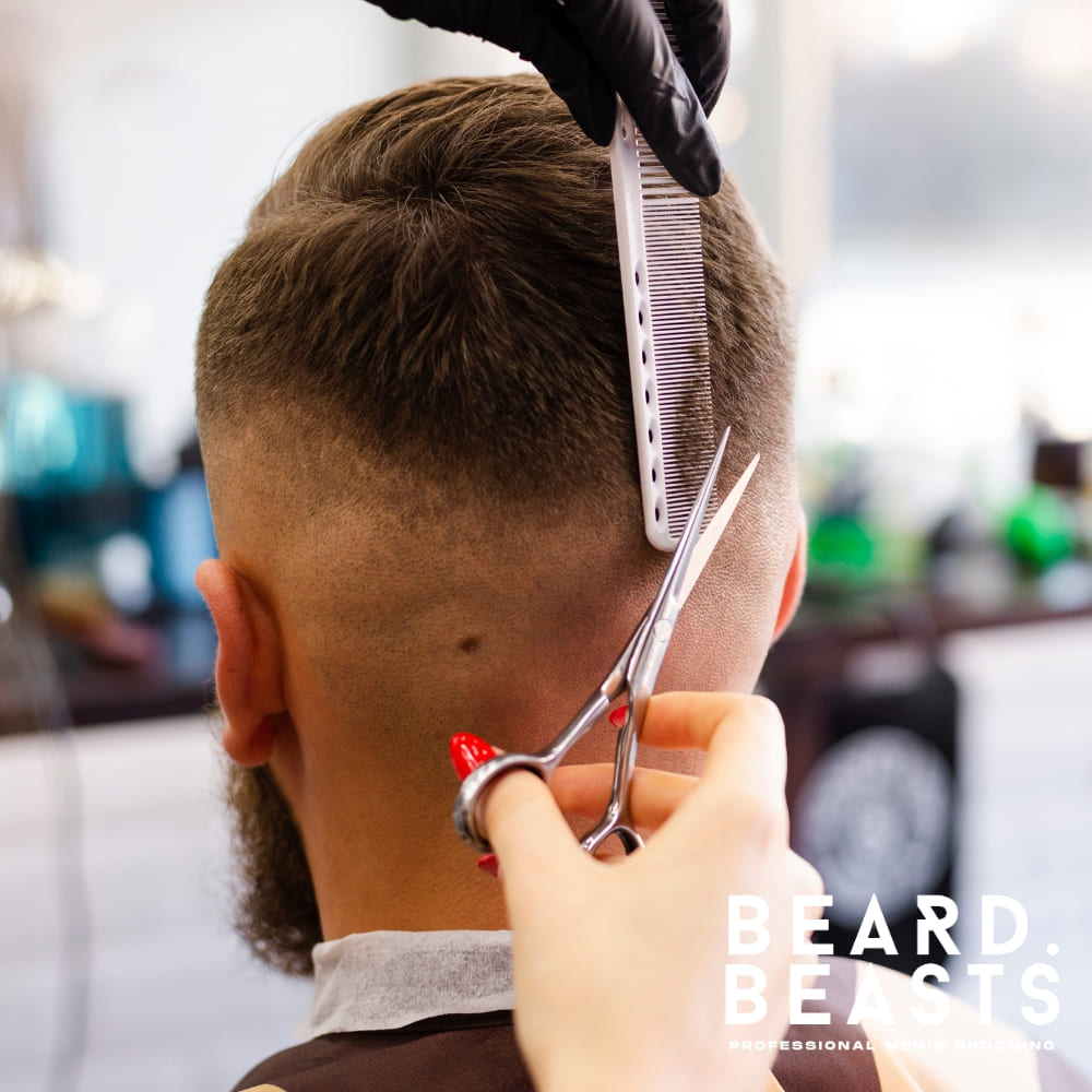 A detailed image of a barber skillfully working on a haircut. The client is seated, with the back of his head clearly visible, showcasing a gradual fade blending smoothly into the skin. The barber uses scissors and a comb to refine the top transition, ensuring precision and a clean finish. The client sports a neatly trimmed beard that complements the fade, creating a polished and well-groomed look. The background features a professional barbershop setting with tools and equipment, reinforcing the high-quality grooming experience.