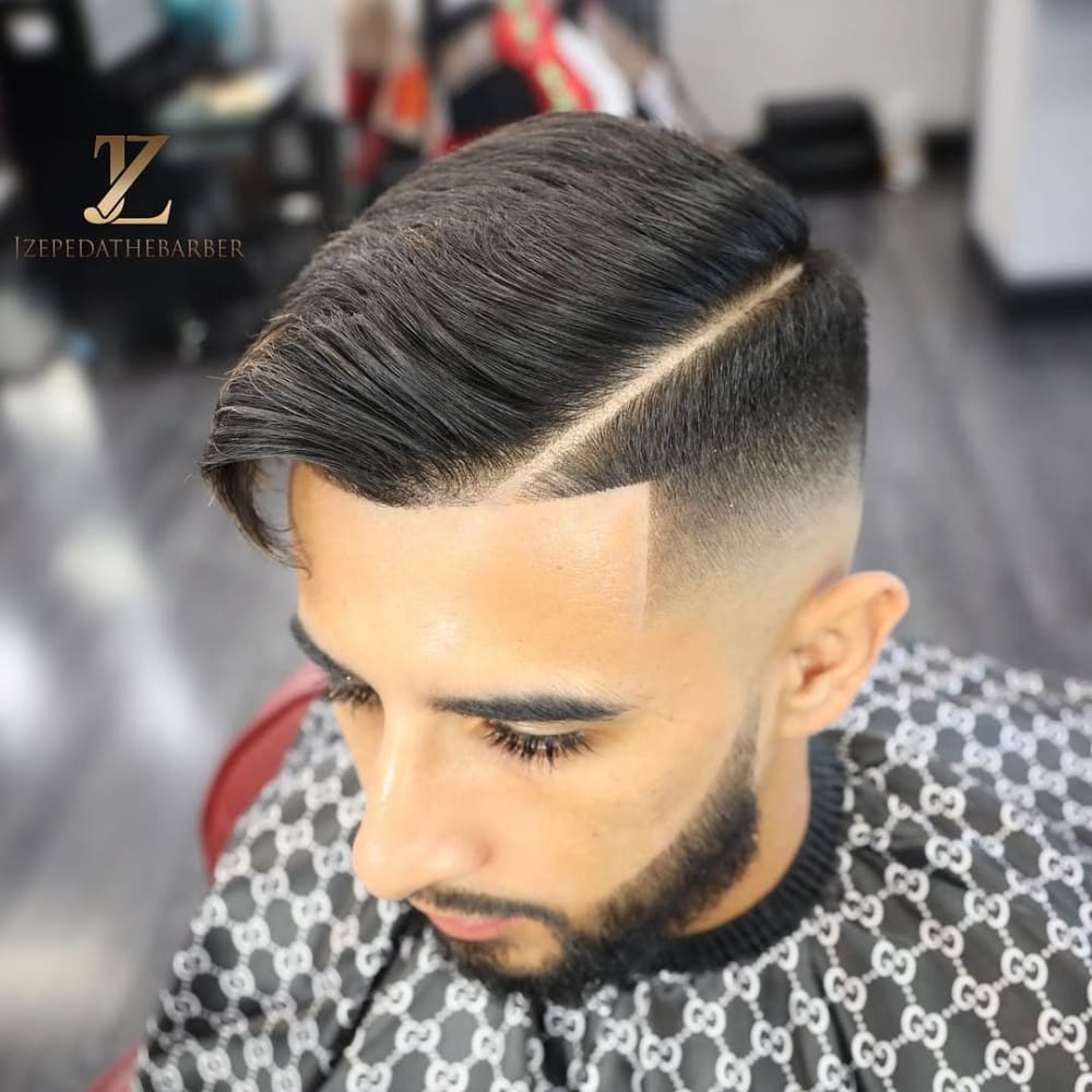 A man with a sharp mid fade comb over haircut, featuring a defined hard part and clean edges, paired with a neatly trimmed beard, seated in a barbershop.