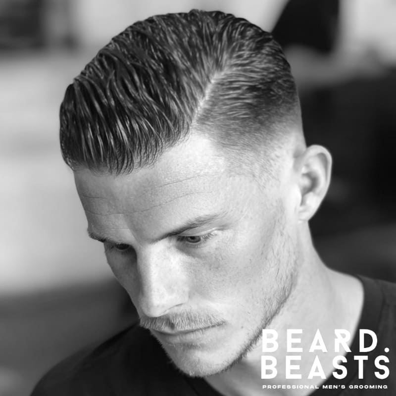 Man with a classic slicked back side part haircut—defined side part, high-shine finish, and neatly blended low fade for a polished, professional look.