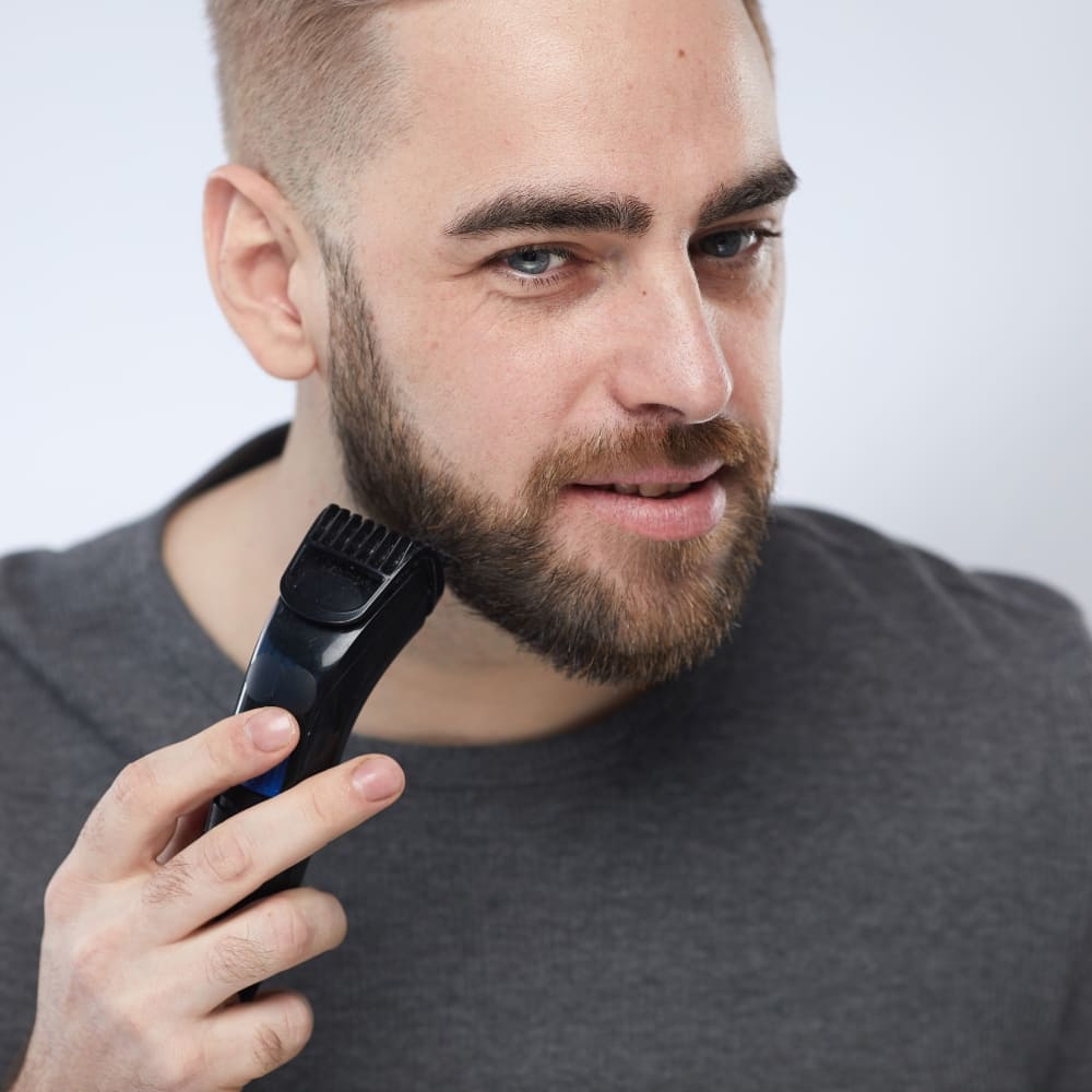 A well-groomed man trimming his beard with an electric trimmer, shaping a short boxed beard for a clean and structured look. His beard is evenly trimmed, maintaining a well-defined and polished appearance. The sharp cheek lines and clean neckline enhance facial symmetry, creating a professional and stylish look. His fade haircut complements the beard, adding to the modern aesthetic. The neutral background and soft lighting emphasize the grooming process, making this a great representation of beard maintenance and self-care.