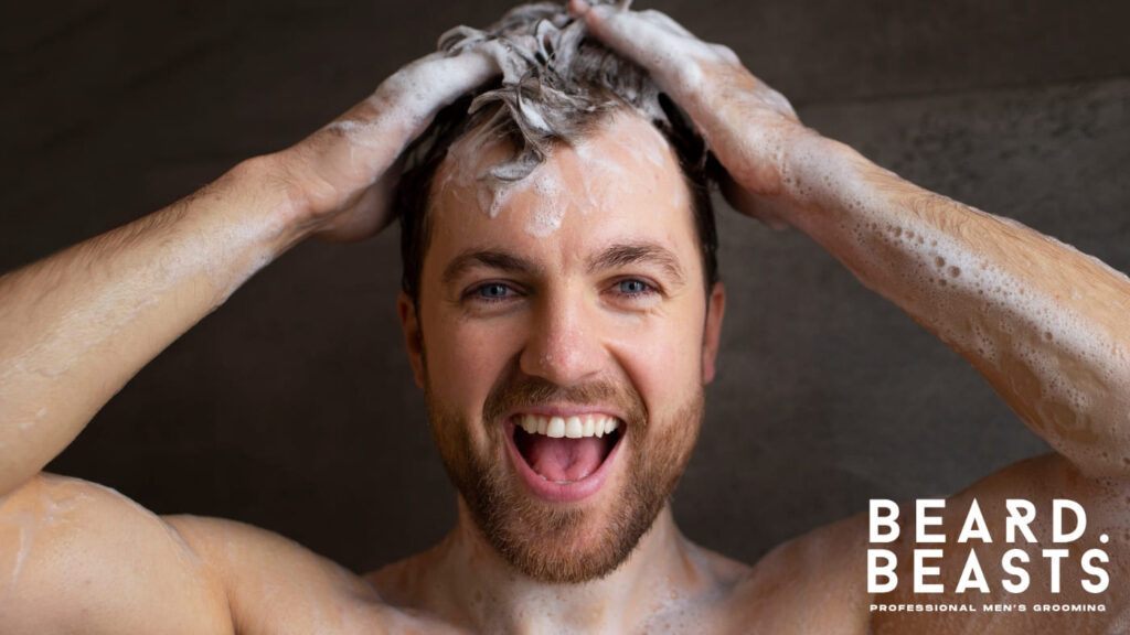 A bearded man washing his hair with shampoo, creating a rich lather, showing how to get pomade out of your hair with the right cleansing routine.