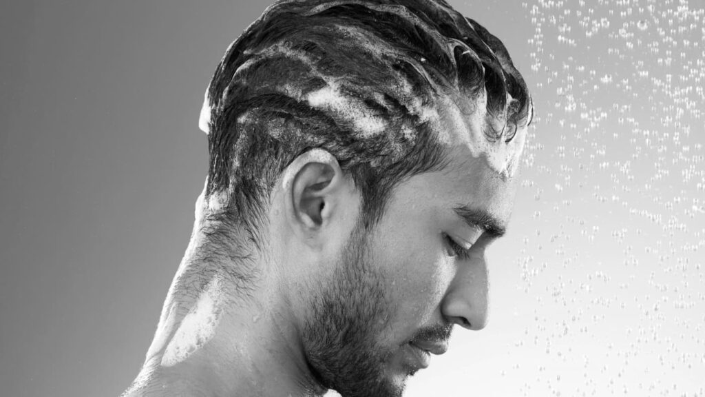 Man washing styling powder out of his hair under running water, with shampoo lather visible. A grooming routine for clean and healthy hair.