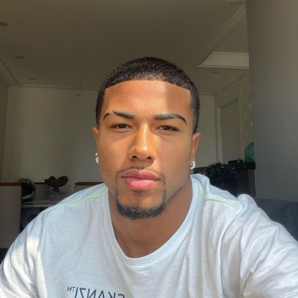 A young man with a neatly trimmed beard and a fresh fade haircut sits in a well-lit room, illuminated by natural sunlight. He has a confident expression, well-groomed eyebrows, and a smooth complexion. He is wearing a white T-shirt with subtle text on it and small stud earrings. The background features a modern interior with neutral tones, including a table with decorative items and a partially visible hallway. His well-defined facial features and sharp grooming give him a stylish and polished appearance.