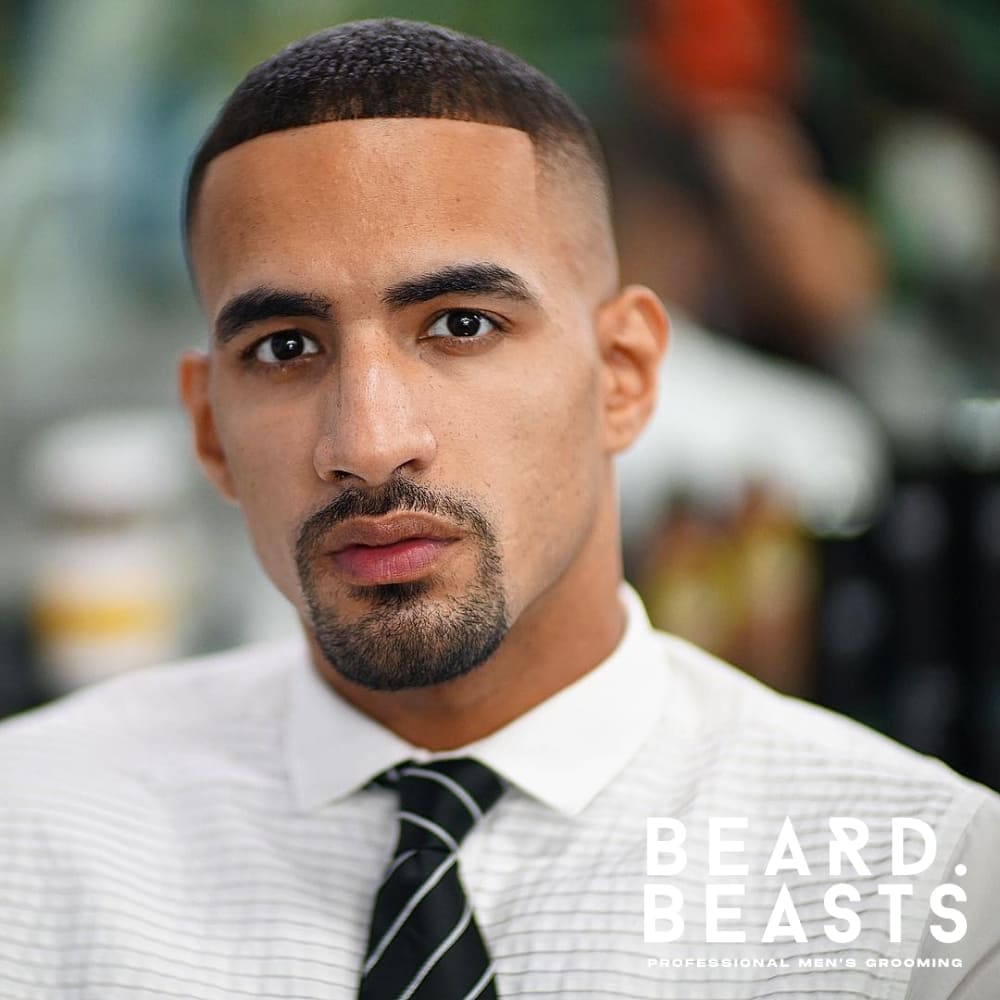 A well-groomed circle beard on a man with a triangle face shape, featuring a wide jawline and a narrow forehead. The beard is neatly trimmed, connecting a defined mustache to a rounded goatee, while the cheeks remain clean-shaven for a sharp, polished look. His faded haircut blends seamlessly with the beard, creating a balanced and professional appearance.