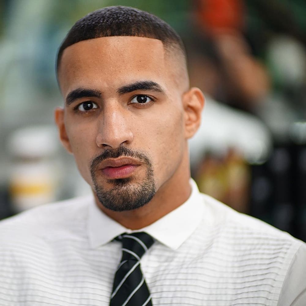 A well-groomed man with a classic circle beard style, featuring a neatly trimmed mustache connected to a rounded goatee. He has a clean-shaven look on the cheeks and jawline, highlighting the sharp definition of his beard. His hair is styled in a fresh fade haircut, complementing his polished appearance. He wears a crisp white dress shirt with a striped black tie, exuding a professional and sophisticated style. The blurred background suggests a barbershop or grooming environment, adding to the refined and stylish aesthetic.