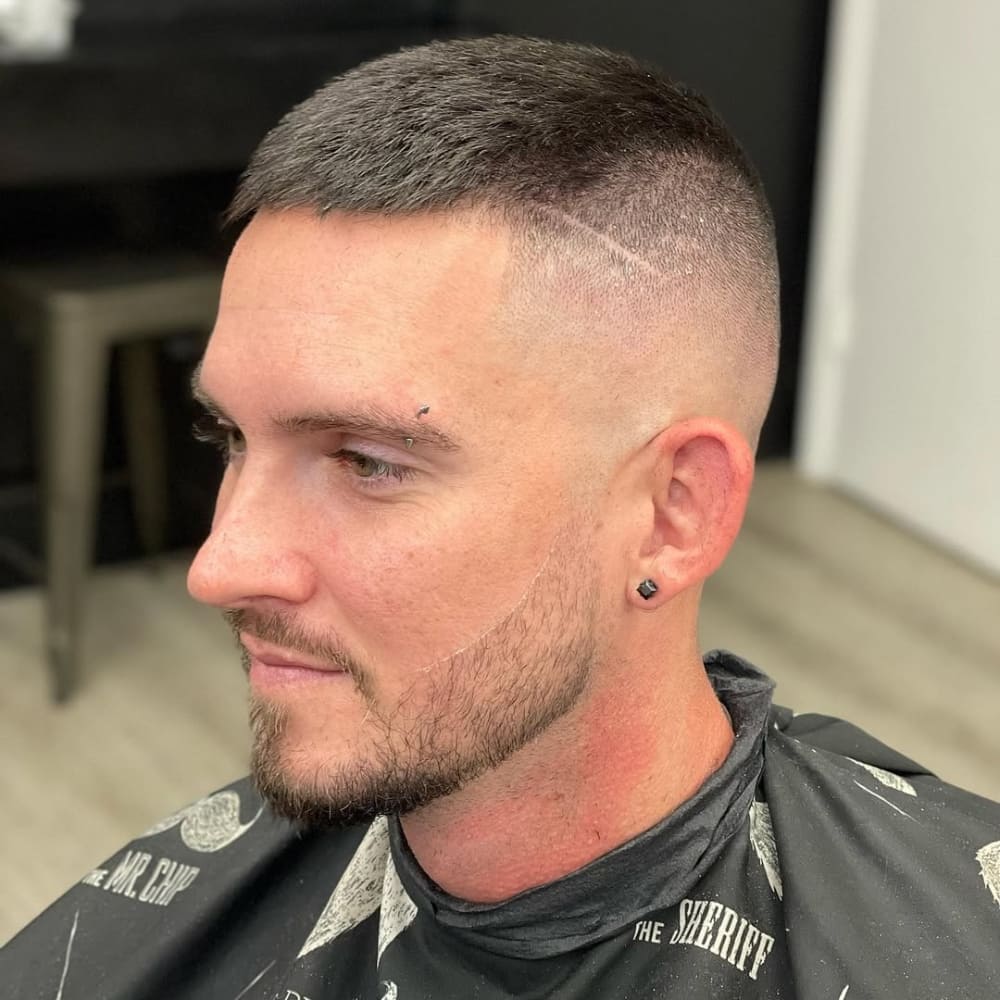 A close-up of a man with a high and tight haircut, featuring an ultra-short fade on the sides and back with a slightly longer top. The fade transitions sharply into the top, creating a clean and structured look, often associated with military-inspired styles. A subtle hair design line is shaved into the fade for added detail. The man has well-groomed facial hair, a neatly trimmed beard, and a confident expression, sitting in a barber chair with a cape draped over him. The background is blurred, emphasizing the haircut.