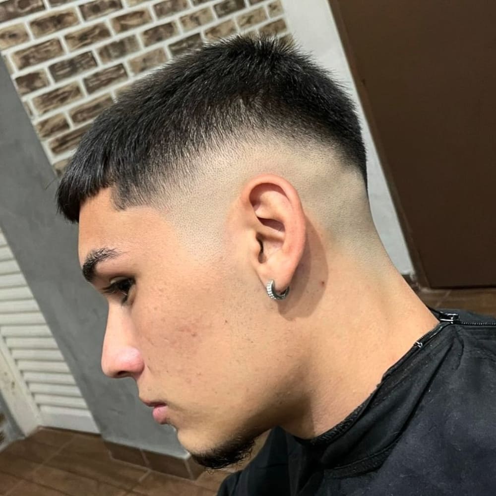 A close-up side profile of a man with a high fade haircut, showcasing a sharp transition from longer hair on top to completely shaved sides. The haircut emphasizes clean lines and a bold, high-contrast look. The man is wearing an earring and a barber cape, seated in a barbershop with a brick and neutral-toned interior.