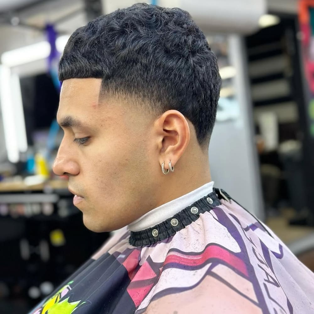 A close-up side profile of a man with a high taper haircut, featuring a clean and gradual transition from longer hair on top to shorter hair near the sides. The haircut highlights neatly defined edges and a slightly textured top, complementing the overall sharp and polished look. The man is wearing earrings and a barber cape, sitting in a well-lit barbershop.