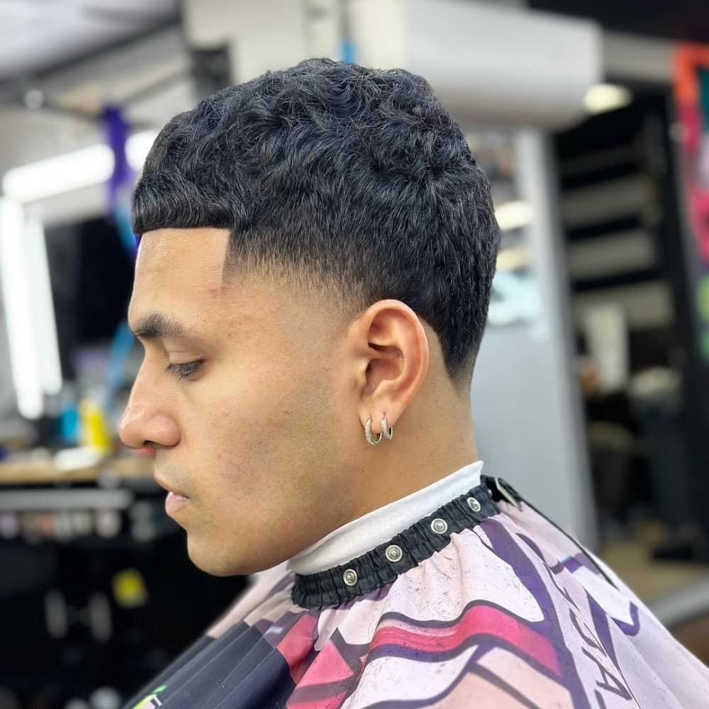 A side profile of a man with a high taper haircut, featuring a sharp fade starting near the temples, paired with short textured curls and hoop earrings.
