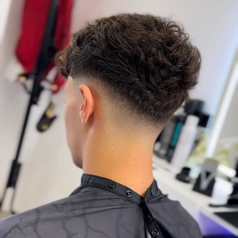 A back view of a man with a clean and sharp low fade haircut, highlighting a smooth gradient from thick, textured hair on top to a skin-close fade near the neckline. The fade begins just above the ears, creating a bold and defined contrast that emphasizes the precision of the haircut. The curly texture on top adds volume and dimension, while the fade provides a polished and modern finish. The image is set in a barber shop, with professional styling tools and products visible in the background, enhancing the professional quality of the haircut.