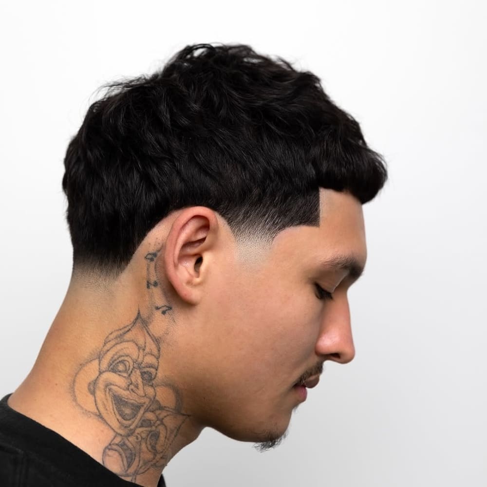 A side profile of a man with a clean and precise low taper haircut, showcasing a gradual transition from longer hair at the top to shorter hair near the neckline. The haircut emphasizes a sharp, defined edge around the ear and neckline for a polished look. The man's hair is textured on top, and he has a visible tattoo on his neck, adding an edgy and unique touch to his overall style. The soft fade blends seamlessly with his natural hair texture, highlighting the professional detailing of the haircut.