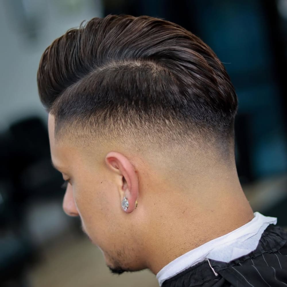 A man with a sleek mid fade comb over hairstyle, featuring a clean fade that transitions smoothly to longer, styled hair on top, photographed in a barbershop setting.