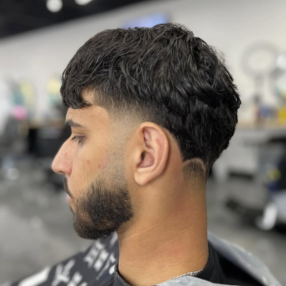 A man with a mid taper haircut, featuring a gradual and natural blend from longer textured hair on top to shorter sides. The taper maintains clean lines around the edges, complementing his beard for a sharp and balanced look. The haircut highlights versatility, offering a modern yet professional style.