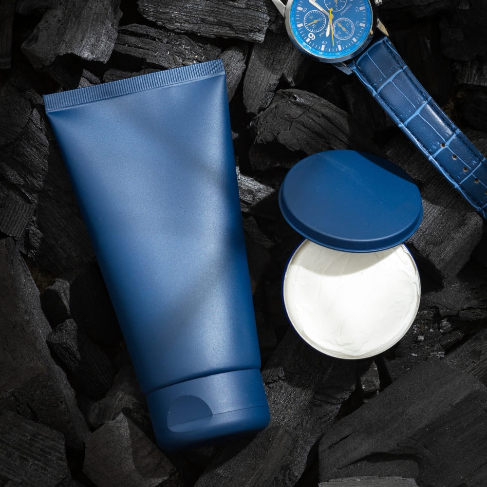 A flat-lay image featuring a blue tube and an open container of men's hair styling cream, placed on dark charcoal pieces, alongside a stylish blue wristwatch with a leather strap, evoking a sleek and modern grooming aesthetic.