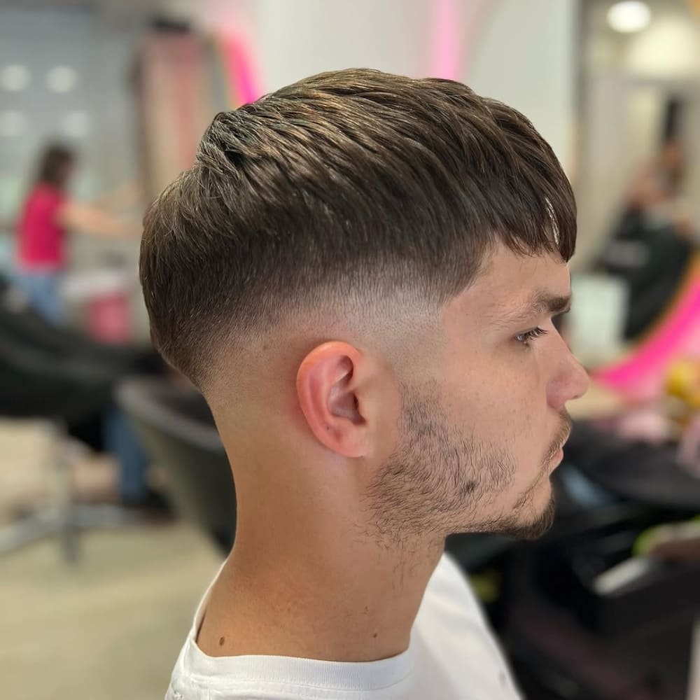 A side profile of a man showcasing a mid skin fade with a French crop hairstyle. The fade begins at the mid-point of the head, transitioning smoothly into the skin for a sharp and clean contrast. The top features a short, textured fringe styled forward, creating the signature look of a French crop. The hair is neatly blended, highlighting the precision of the fade and the balance between the top and sides. The setting is a modern salon, with soft lighting and background activity that adds context to the fresh haircut.