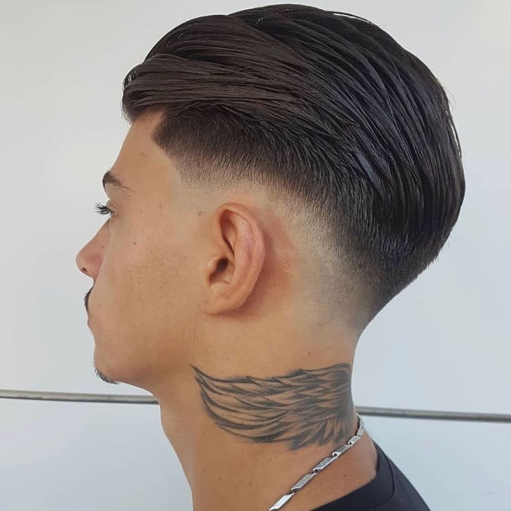 A close-up side profile of a man with a mid skin fade hairstyle paired with a sleek, slicked-back top. The fade begins at the mid-point of the head and blends seamlessly into the skin, creating a sharp and clean look. His straight hair is styled smoothly back for a polished and professional appearance. The image also features a detailed wing tattoo on the back of the neck, adding a bold personal touch.