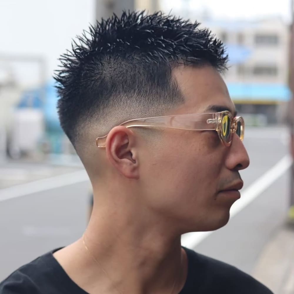 A side profile of a man sporting a mid skin fade with a spiky top hairstyle. The fade begins at the mid-point of the head and blends smoothly into the skin, creating a clean and sharp contrast with the textured top. The hair on top is styled into bold, defined spikes for a modern, edgy look. The man is wearing transparent sunglasses, adding a touch of personality to the overall style. The outdoor setting provides a natural backdrop, emphasizing the fresh and trendy haircut.