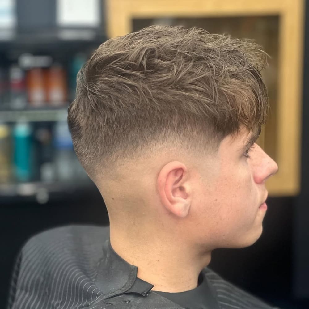 A side profile of a man with a mid skin fade hairstyle complemented by a textured crop. The fade starts at the mid-point of the head and transitions smoothly into the skin, creating a clean and sharp contrast. The top features tousled, choppy layers, adding volume and texture for a relaxed yet stylish appearance. The setting is a barbershop, with grooming products visible in the background, highlighting the fresh and professional haircut.