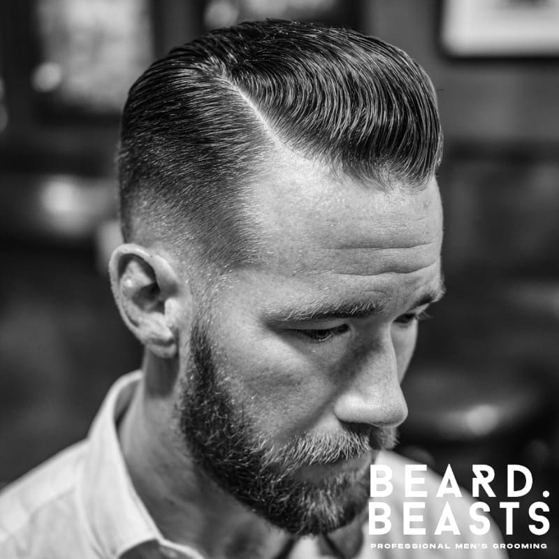 Man with a slicked back side part and low fade—clean taper around the ears, sharp side part, and polished finish with beard.