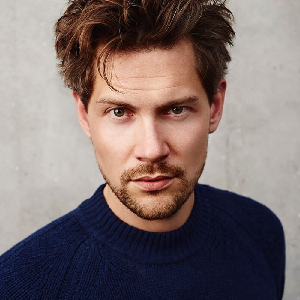 A young man with tousled brown hair and a well-groomed short facial hair poses against a neutral concrete background. His facial hair is light and even, creating a subtle yet defined look that enhances his sharp jawline. He wears a navy blue knitted sweater, adding a casual yet stylish touch to his appearance. His green eyes and confident expression give him a modern and approachable look. The soft lighting highlights his facial features and the natural texture of his facial hair.