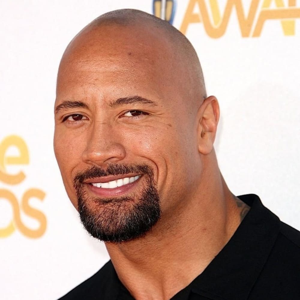 A bald man with a well-groomed, thick beard smiles confidently at an awards event. His facial hair is neatly trimmed into a thick circle beard, with a fully connected mustache and a rounded goatee that enhances his strong jawline. He is wearing a black collared shirt, complementing his polished and professional appearance. The bright lighting highlights his smooth complexion and the clean definition of his beard. The blurred background features event signage, indicating a red carpet or celebrity gathering setting.