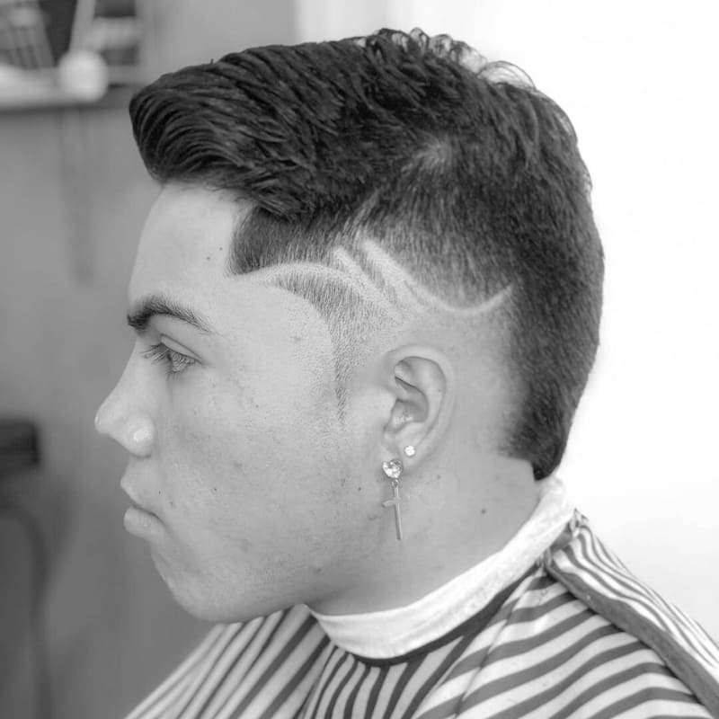A young man with a burst fade haircut featuring intricate hair designs shaved into the sides. The fade seamlessly blends into the longer textured top, creating a bold and stylish contrast. The shaved design adds a unique, artistic touch, enhancing the modern and edgy look. He is wearing multiple earrings, including a dangling cross, and a striped barber cape, suggesting he is in the middle of a fresh haircut session. The black-and-white composition emphasizes the sharpness of the fade and the precision of the design.