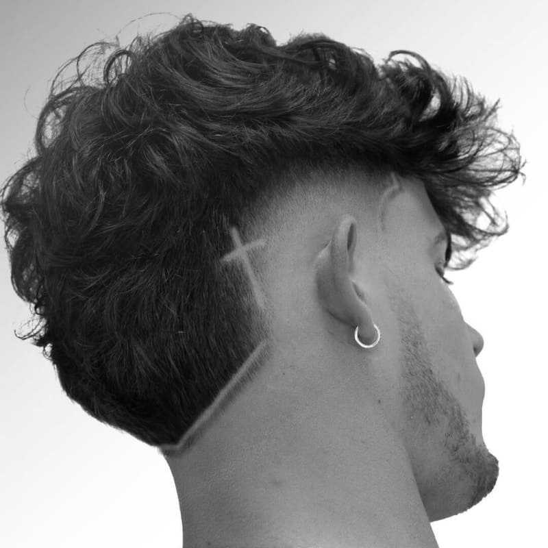 A stylish Burst Undercut Fade featuring a sharp design etched into the fade. The haircut showcases a high-contrast transition with voluminous, curly hair on top left disconnected from the faded sides, creating a bold undercut effect. The fade is clean and precise, curving around the ear, with a distinct cross design adding a unique touch. The subject wears a silver hoop earring and a neatly trimmed beard, complementing the edgy, modern look.