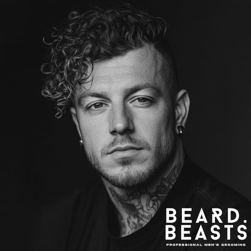 A stylish man with a curly comb over haircut, featuring a voluminous, side-swept curly top with a sharp undercut for contrast. His natural curls add texture and movement, giving the hairstyle a bold yet effortless appeal. A well-groomed beard and intricate neck tattoo enhance his edgy and modern look. Dressed in a black t-shirt against a dark background, the monochrome composition emphasizes the depth and structure of his haircut. The