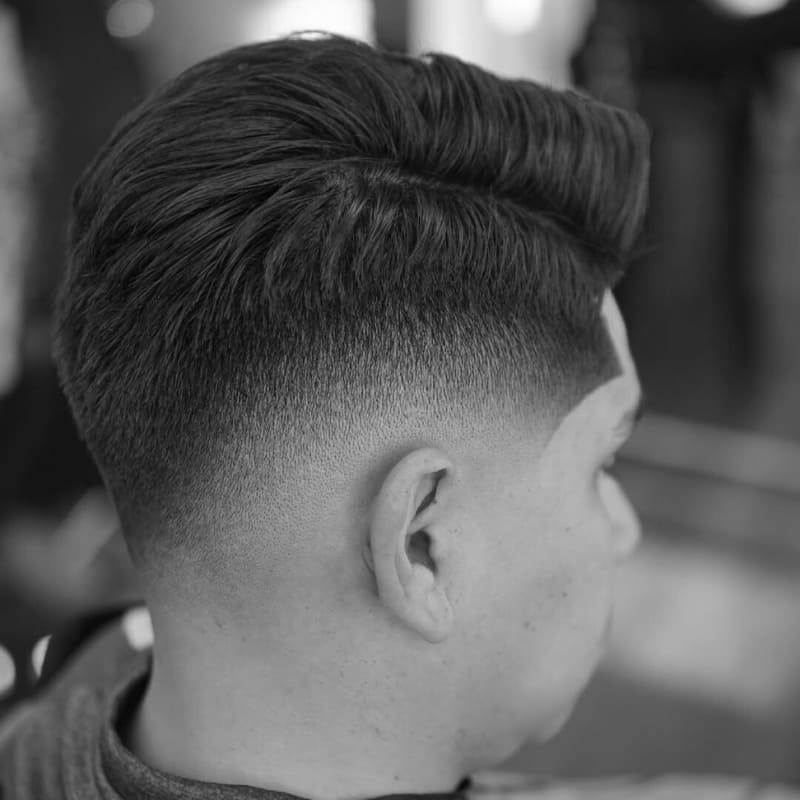A close-up view of a drop fade haircut, showcasing a seamless transition from the neatly faded sides to the longer, voluminous top. The fade curves naturally around the head, creating a smooth and stylish contrast. The hair on top is textured and styled for a clean yet modern appearance. The sharp lineup along the temple enhances the precision of the cut. The black-and-white composition highlights the depth and detail of the fade, emphasizing the craftsmanship of the haircut.