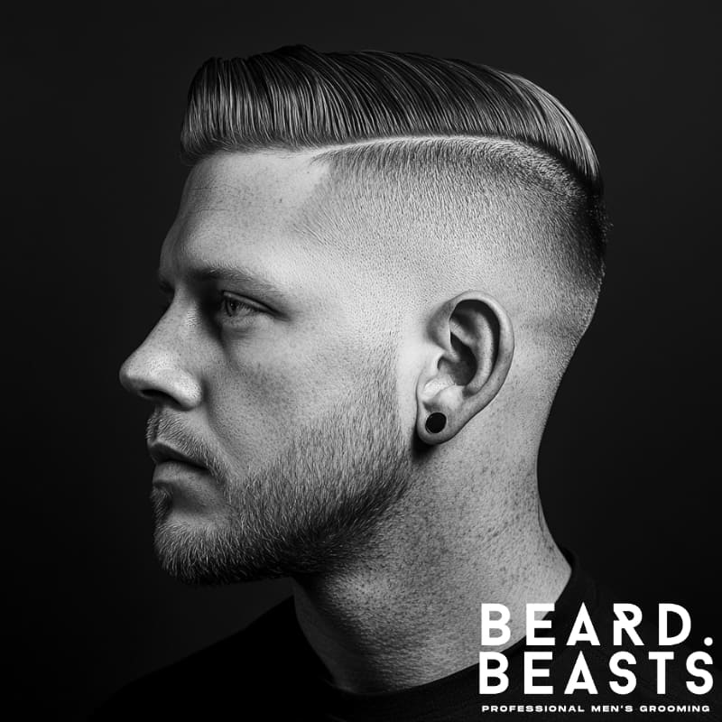 A sharp and modern drop hairstyle featuring a precisely styled hard part. The sleek, combed-back top contrasts with the smoothly faded sides that follow the natural curve of the head, creating a clean, structured look. The well-groomed beard complements the hairstyle, adding a masculine and polished edge. The black-and-white aesthetic enhances the sharp definition of the cut, making it stand out as a bold and stylish choice. The Beard Beasts logo in the corner reinforces the brand's focus on professional men's grooming.