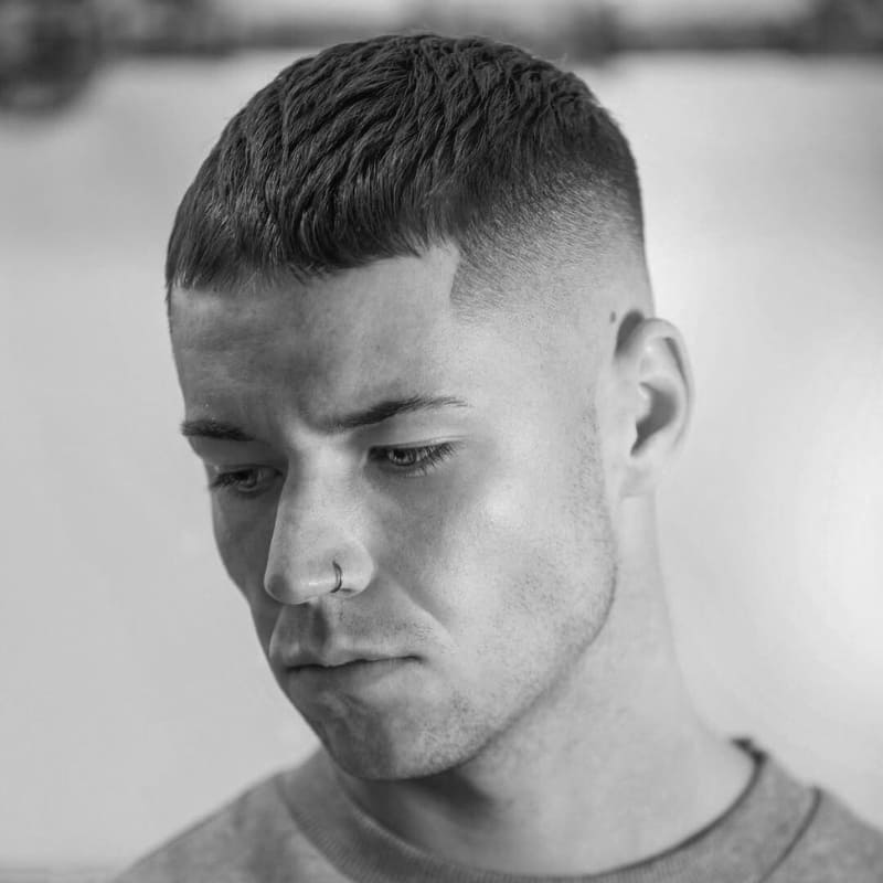 A black-and-white portrait of a young man with a high fade haircut, one of the boldest types of fades for men. The fade starts high on the sides, creating a sharp contrast with the short, textured crop on top. His hairline is precisely shaped, enhancing the structured and modern look. The smooth gradient of the fade highlights expert barbering techniques. This clean and edgy style is ideal for those who prefer a sharp, well-defined appearance.