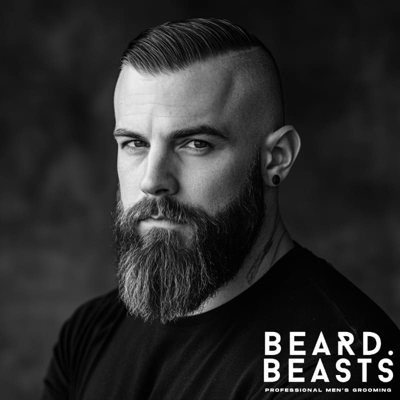 Black-and-white portrait of a man with a high and tight comb over and full beard style. His hair is sharply parted and combed to one side with a clean, high fade on the sides. The long, thick beard is well-groomed, creating a bold contrast with the short hairstyle. This look combines sharp structure with rugged facial hair for a strong, masculine appearance.