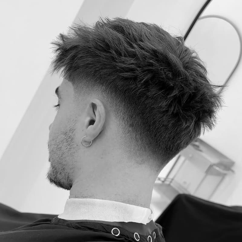 A black-and-white photograph of a young man with a low fade haircut and a textured, messy top. The fade starts just above the ears, creating a smooth and subtle transition that blends seamlessly into the longer hair. His neckline is cleanly tapered, enhancing the overall polished look. He is wearing a small hoop earring, adding a stylish touch to his appearance. The image is taken from behind in a barbershop setting, highlighting the precise detailing of the haircut.