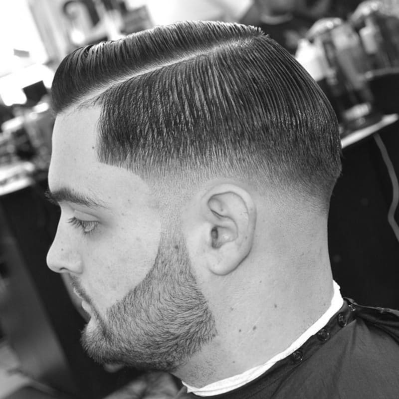 A black-and-white close-up of a man with a low fade haircut, featuring a precise fade that gradually tapers down the sides. The top is styled sleek and neatly parted, creating a sharp contrast with the clean fade. His well-groomed beard is edged to perfection, complementing the structured look. The haircut has a polished finish, achieved with a styling product for a smooth and controlled appearance. The barbershop background adds authenticity, emphasizing the fresh, professional cut.
