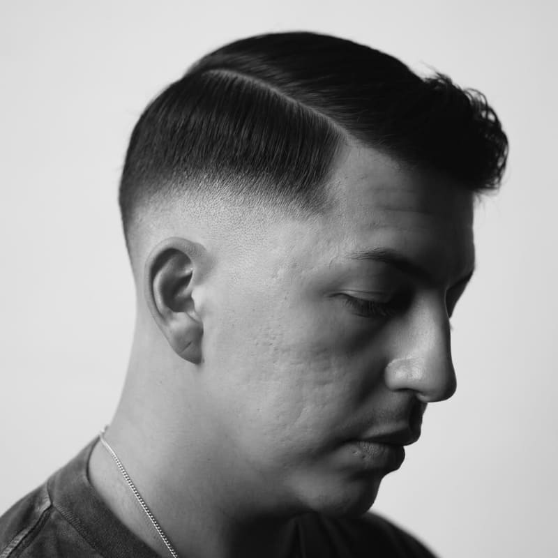A black-and-white close-up portrait of a man with a mid fade haircut, featuring a smooth transition from the neatly faded sides to the longer, styled top. The sharp parting adds definition, enhancing the structured and polished look. The fade is clean and precise, creating a seamless blend between the shaved area and the textured upper section. His expression is calm and introspective, with soft lighting highlighting the contours of his face. The monochrome composition emphasizes the sharp details of the haircut and the overall grooming precision.