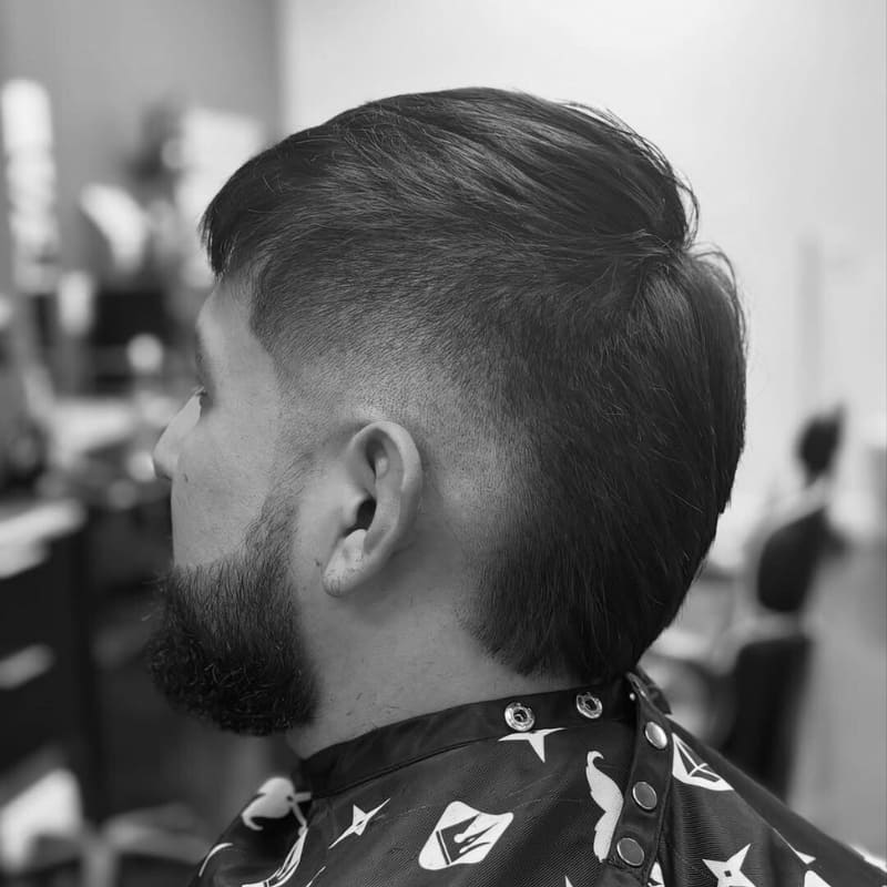 A stylish modern haircut featuring a mohawk-inspired look with neatly tapered sides. The top is left long and combed back, creating a smooth contrast with the closely trimmed sides. The subject has a well-groomed beard that enhances the sharp and structured appearance. The photo is taken in a professional barbershop setting, highlighting the precision of the haircut. The black and white filter adds a classic, edgy feel to the image.