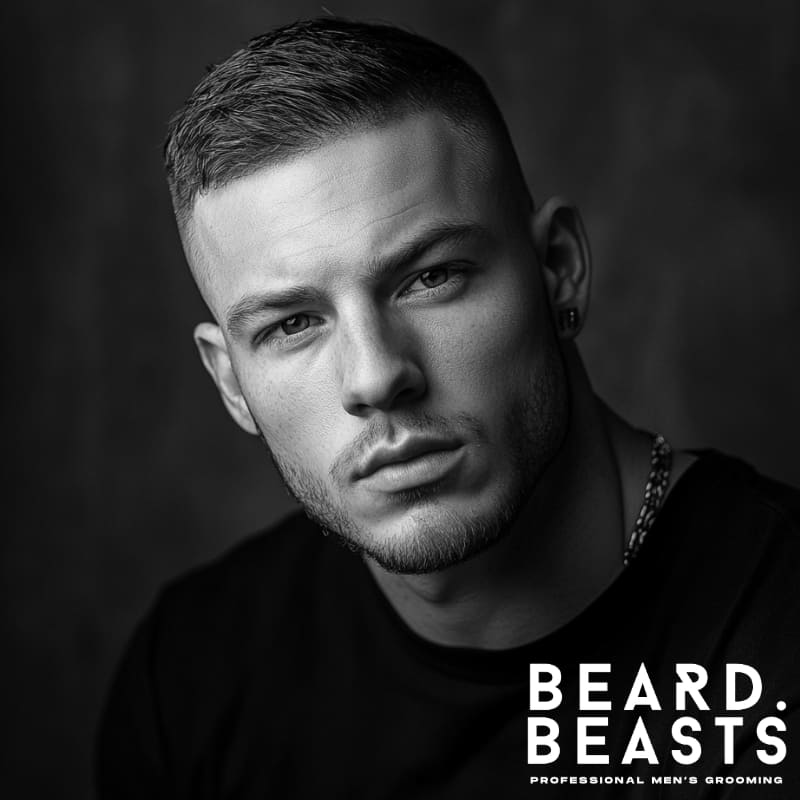 A confident man with a short hair comb over, featuring a neatly styled top with a subtle side-swept texture and a clean, low fade on the sides. His sharp facial features are complemented by a well-groomed light beard, adding to his modern and masculine look. Wearing a fitted black t-shirt and a stylish chain necklace, he exudes effortless confidence. The monochrome composition enhances the contrast and detail, emphasizing the structure of the haircut and facial definition. The