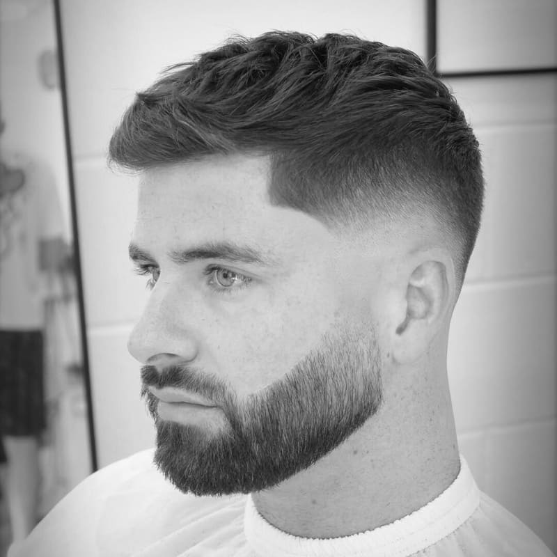 A black-and-white photograph of a young man with a skin fade haircut, one of the sharpest types of fades for men. The fade starts high on the sides and gradually blends down to the skin, creating a clean and seamless transition. His textured hair on top adds volume, while his precisely shaped beard enhances the overall structured look. The sharp hairline and smooth fade highlight expert barbering techniques. This style offers a modern, polished appearance perfect for those who prefer a crisp, well-defined fade.