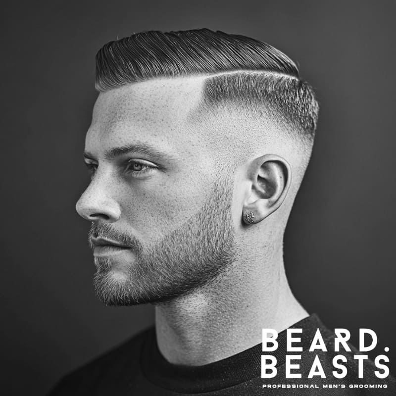A black-and-white side-profile portrait of a man with a skin fade comb over undercut, featuring a precise hard part and a seamless fade that blends down to the skin. His hair is styled sleek and neatly combed to the side, emphasizing the clean contrast between the sharp top and closely shaved sides. A well-groomed short beard complements the hairstyle, adding balance to his facial features. His expression is calm and confident, with a blurred background that keeps the focus on his haircut and grooming details.