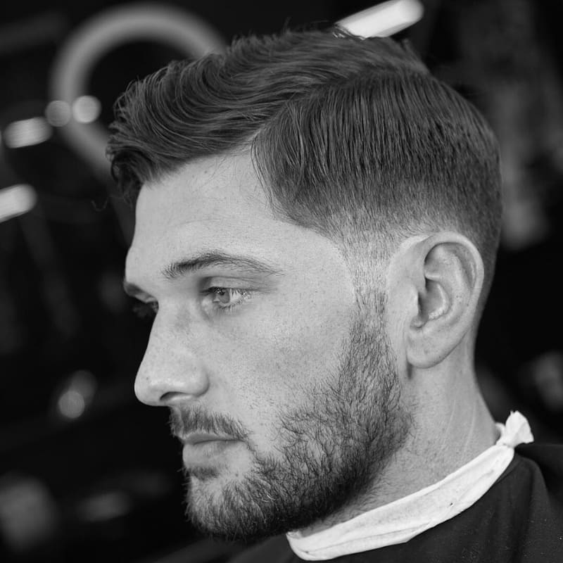 A black-and-white close-up portrait of a man with a taper fade haircut, featuring a smooth gradient from the neatly faded sides to the longer, textured top. His hair is styled with a natural, slightly tousled look, complementing his well-groomed beard. The precise fade creates a sharp yet subtle contrast, enhancing the overall clean and polished appearance. Captured in a barbershop setting, the blurred background highlights the fresh, professional cut. The monochrome composition emphasizes the depth and definition of the fade and hair texture.
