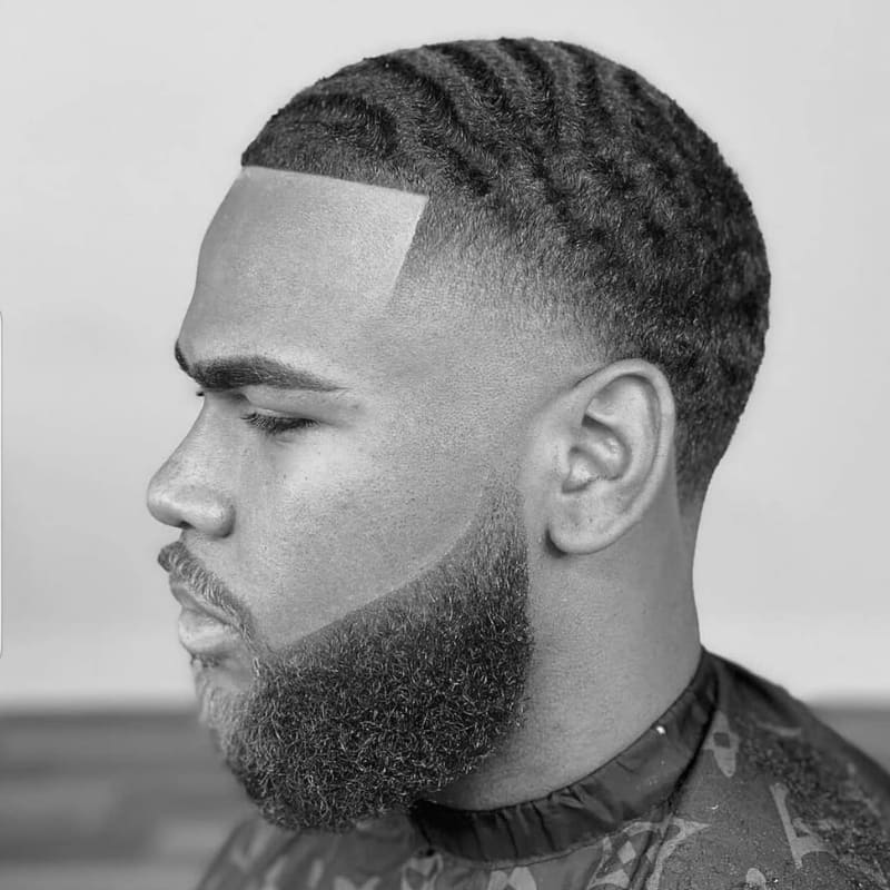A black-and-white photograph of a young man with a temple fade haircut, one of the most popular types of fades for men. The fade is precisely blended at the temples, creating a sharp and clean transition into his neatly styled 360 waves. His hairline is expertly shaped, and his full, well-groomed beard complements the fade for a polished look. The contrast between the sharp fade and the textured waves highlights the precision of the cut. The image captures a professional barbershop-style finish, emphasizing modern men's grooming trends.